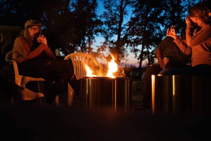 Three people enjoying the solo flame fire pit outdoors with chopped firewood nearby.